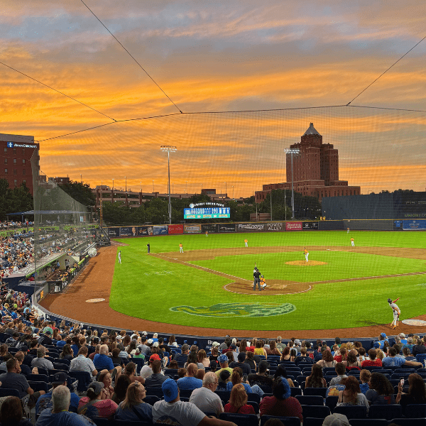 A high-angle view from behind home plate of a baseball game at 7 17 Credit Union Park during a vibrant sunset over downtown Akron, with spectators filling the stands.